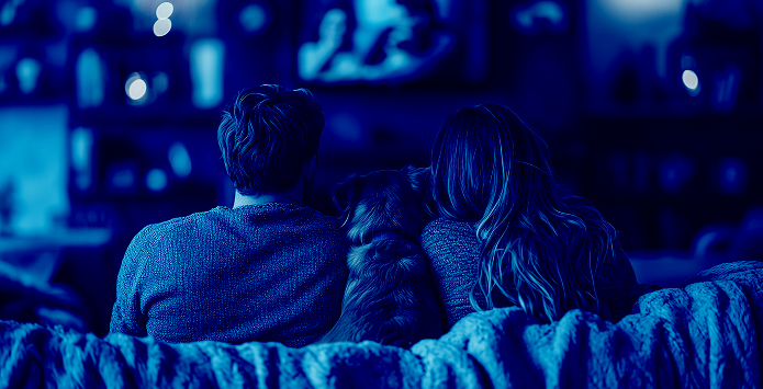 a back view of a man and a woman sitting on a sofa with a dog between them, watching a television screen in a dimly lit room, shown with a monochromatic blue filter to represent shared viewing experiences.
