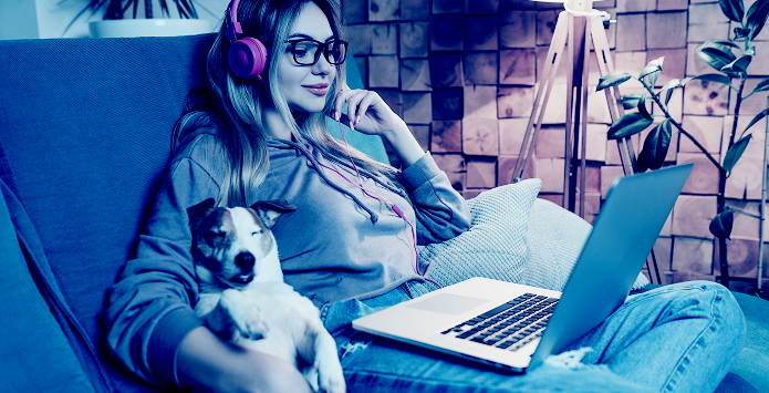A woman wearing pink headphones and glasses sits on a couch using a laptop with a small dog resting beside her, shown through a blue color filter.