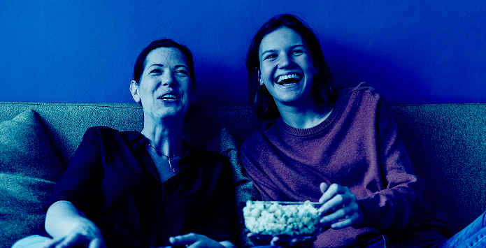 Two women sitting on a sofa, laughing and sharing a bowl of popcorn while watching a screen, presented with a monochromatic blue colour filter to illustrate household engagement.