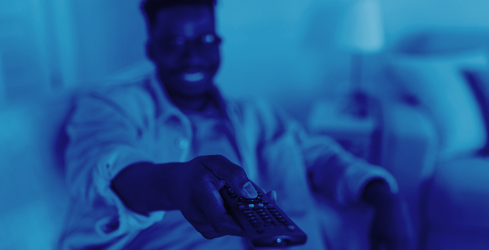 A man sitting on a sofa in a dimly lit room, holding a TV remote and smiling toward a screen, shown through a monochromatic blue colour filter.