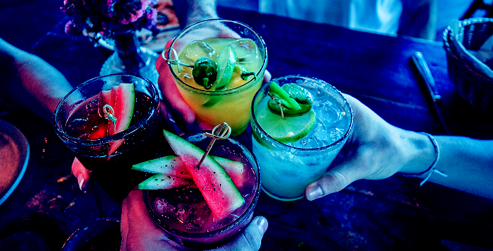 An overhead, close-up shot of four hands holding vibrant, garnished cocktails in a toast over a dark table. the image uses moody, neon lighting with deep blues and pinks, featuring drinks with watermelon and citrus slices.