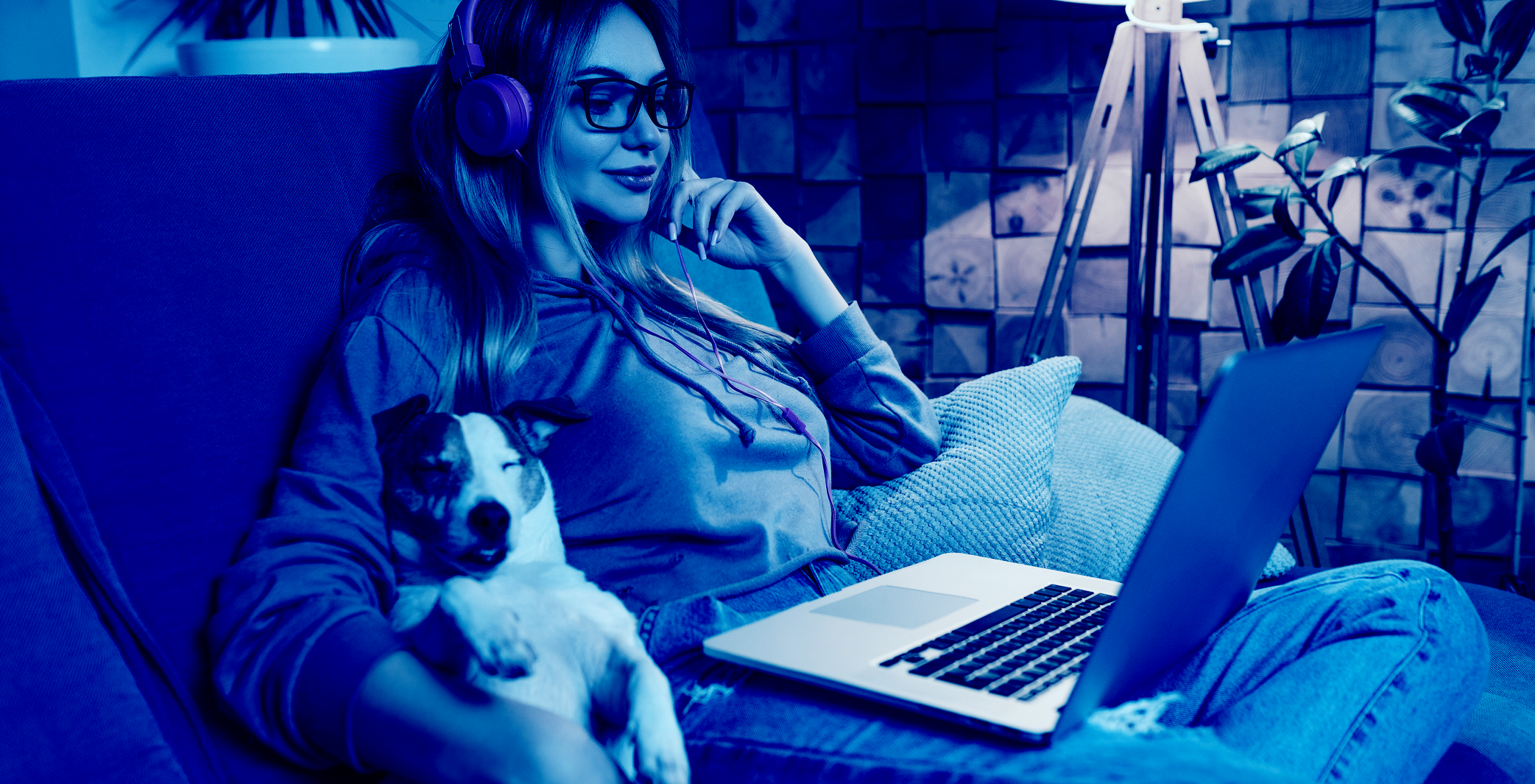 A woman wearing pink headphones and glasses sits on a couch using a laptop with a small dog resting beside her, shown through a blue color filter.