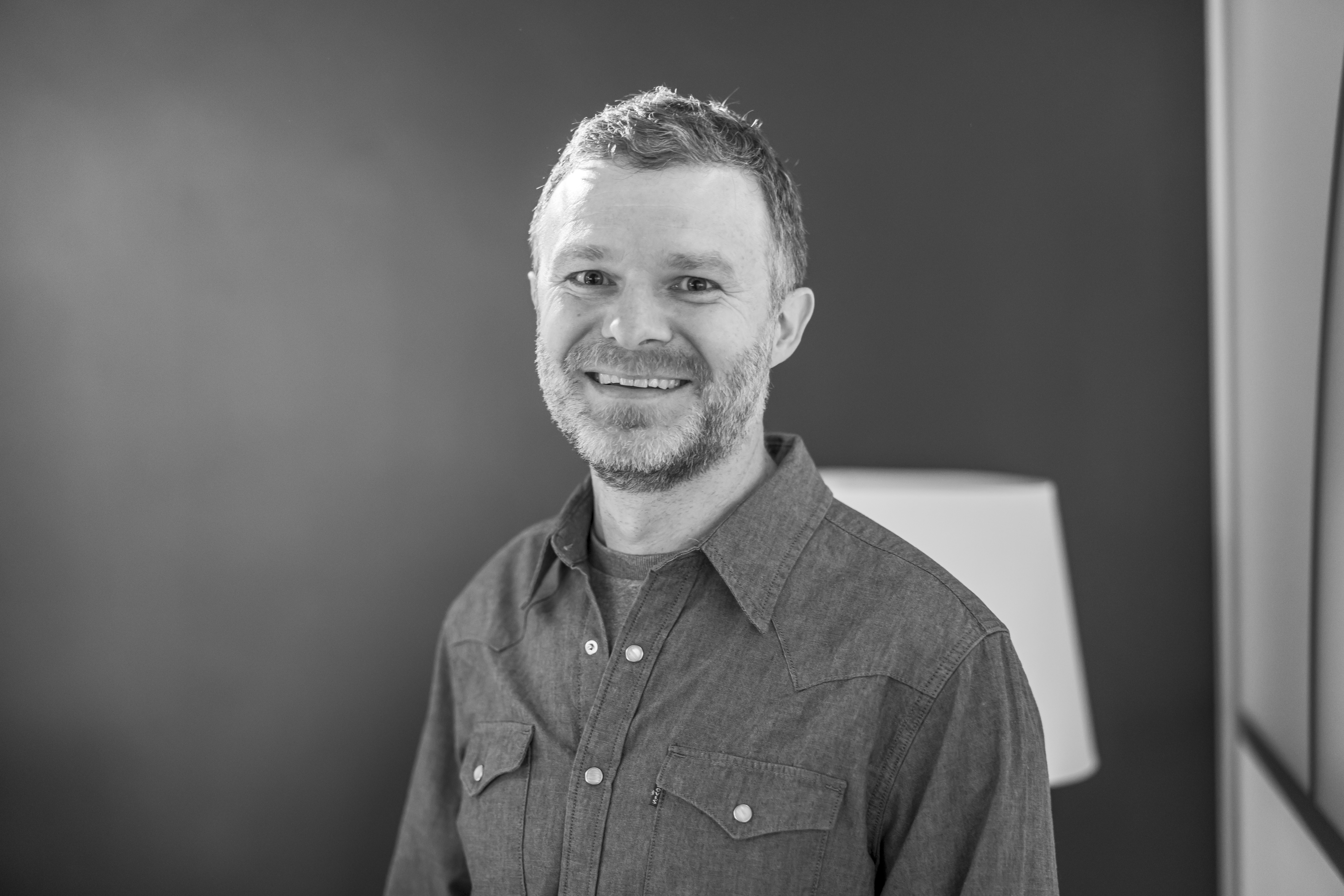 "A black and white head-and-shoulders portrait of Dan (Covatic's CPO), a smiling man with a short beard, wearing a dark denim western-style button-up shirt against a blurred office background."